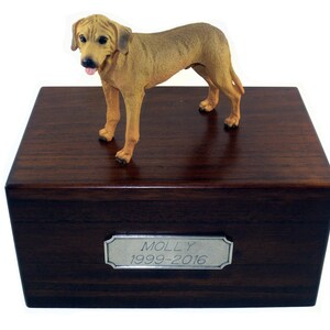 May include: A rectangular dark brown wooden box with a silver-toned metal plaque engraved with the text MOLLY 1999-2016. A tan-colored dog figurine with black ears and a pink tongue is positioned on top of the wooden box lid.