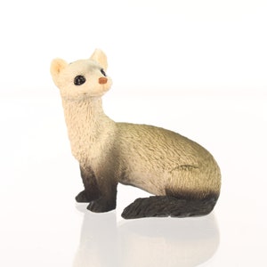 May include: A small, detailed figurine of a ferret. The ferret has a cream-colored head and neck, transitioning to a light brown body and a dark brown tail. It is posed in a seated position, looking upwards.