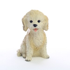 May include: A small, white, fluffy dog figurine with black eyes and a pink tongue sticking out. The figurine is sitting with its legs crossed and its tail curled.