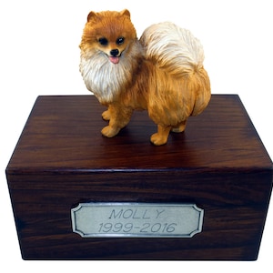 May include: A wooden pet urn with a small, orange and white Pomeranian figurine on top. The urn has a silver plaque with the name "MOLLY" and the dates "1999-2016" engraved on it.