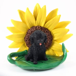 May include: Black miniature poodle figurine sitting on a green leaf with a yellow sunflower behind it.