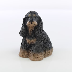 May include: A black and brown ceramic figurine of a cocker spaniel dog sitting with its head tilted down. The figurine has a realistic appearance with detailed fur and facial features.