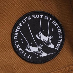 Può includere: Una toppa rotonda nera con testo bianco e un'immagine di scarpe da tip tap. Il testo recita "IF I CAN'T DANCE IT'S NOT MY REVOLUTION". La toppa è cucita su un tessuto marrone.