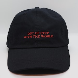 Könnte beinhalten: Eine schwarze Baseballkappe mit rotem Sticktext, der "OUT OF STEP WITH THE WORLD" lautet.