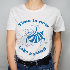 Peut inclure: T-shirt blanc avec un dessin de abeille bleu et le texte "Time is now take a stand".