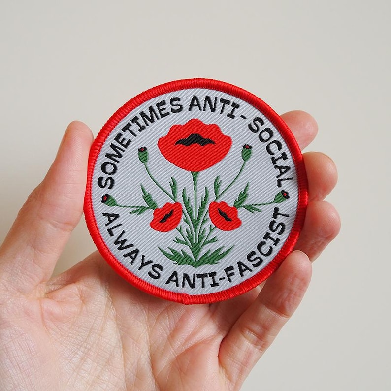 Op de afbeelding: Een geborduurde patch in rood, wit en zwart met de tekst "Sometimes Anti-Social Always Anti-Fascist" en een gestileerde afbeelding van drie rode klaprozen met groene bladeren.