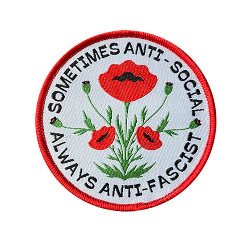 Op de afbeelding: Een witte ronde patch met een rode rand. De patch heeft een ontwerp van drie rode klaprozen met groene stelen en bladeren. De tekst "Sometimes Anti-Social Always Anti-Fascist" staat in een cirkel rond de klaprozen.
