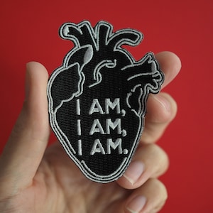 Könnte beinhalten: Schwarzer gestickter Bügel-Patch in Herzform mit dem Text "I AM, I AM, I AM." in weißem Garn.
