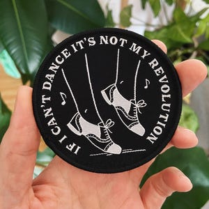 Puede incluir: Parche circular negro con texto blanco y un dibujo de zapatos de baile. El texto dice: "If I can't dance it's not my revolution". También se incluyen notas musicales en el diseño.