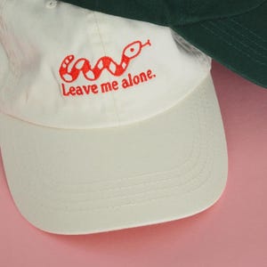 Pode incluir: Boné de beisebol creme com um desenho de cobra vermelha e a frase "Leave me alone". O boné tem uma aba curva e um fecho de fita. Um segundo boné verde é parcialmente visível.