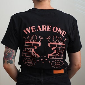 Könnte beinhalten: Schwarzes T-Shirt mit einem rosafarbenen Motiv einer zerbrochenen Mauer aus Ziegeln mit Stacheldraht oben. Der Text "WE ARE ONE" befindet sich über der Mauer. Unter der Mauer befinden sich zwei Schilder mit der Aufschrift "OPEN BORDERS" und "OPEN MINDS".