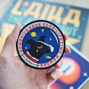 Op de afbeelding: Een ronde patch met de tekst "HOMESICKNESS CLUB LIFETIME MEMBER" rond een blauwe achtergrond met een hondenillustratie. De hond heeft een zwarte kop, een wit gezicht en een enkele traan. De patch heeft ook het woord "LAIKA" in Cyrillisch.