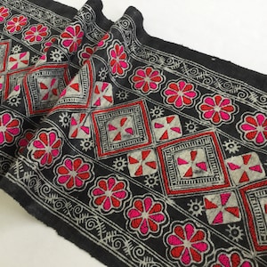 May include: Black fabric trim with a repeating pattern of white and gray geometric shapes and pink and red flowers. The trim is embroidered with a white thread.