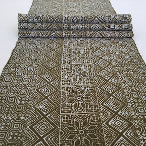 May include: A brown and white patterned fabric table runner with a geometric design. The runner is folded in thirds and has a fringed edge.