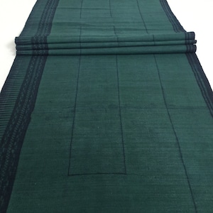 Puede incluir: Un camino de mesa de algodón verde oscuro con un patrón geométrico en un tono más claro de verde. El camino de mesa tiene un borde negro con un patrón repetitivo.