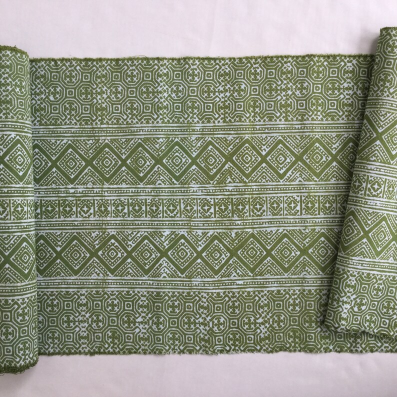 Pu&ograve; includere: Un rotolo di tessuto verde con un motivo geometrico bianco. Il tessuto ha un motivo ripetitivo di diamanti e quadrati.