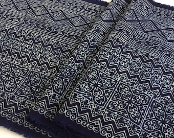 May include: Indigo blue fabric with a repeating white geometric pattern. The design includes diamond shapes, zigzags, and floral motifs. The fabric is folded, showing the detailed pattern and frayed edges.