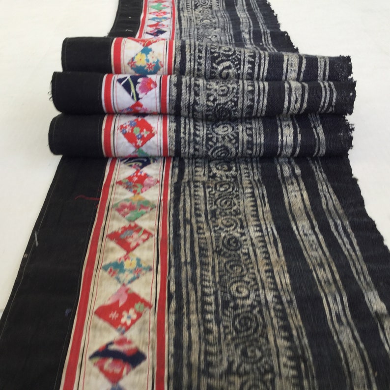 May include: A rolled textile with a black background and a patterned design. The textile features a central band with colorful diamond shapes and a red border. The main section has vertical stripes and decorative patterns in a lighter color.
