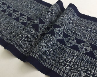 May include: A length of indigo blue fabric with a repeating white geometric pattern. The design features diamond and square shapes with intricate details. The fabric is folded, showing the raw edges and texture of the material.