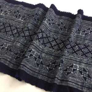May include: Navy blue fabric with a repeating geometric pattern in white. The design includes triangles, diamonds, and lines. The fabric has a fringed edge and is folded, showing the texture and drape of the material.