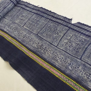 May include: A close-up of a blue and white fabric with a repeating pattern of squares and intricate designs. The fabric has a dark blue border with a colorful embroidered trim.