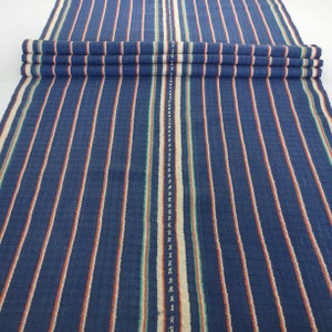 May include: A blue and white striped fabric with thin red and green stripes. The fabric has a woven texture.