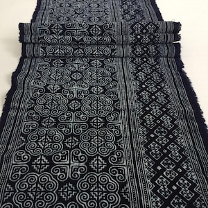 May include: A folded length of indigo fabric with a repeating white pattern. The design features swirling cloud-like shapes and geometric elements. The edges are frayed, and the fabric is likely used for textiles or home decor.