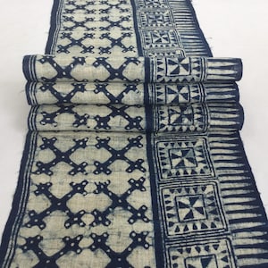 Peut inclure: Textile roulé avec un fond crème et des motifs géométriques bleu marine. Le motif comprend des formes en croix et des motifs carrés. Le tissu semble être du lin ou une fibre naturelle similaire.