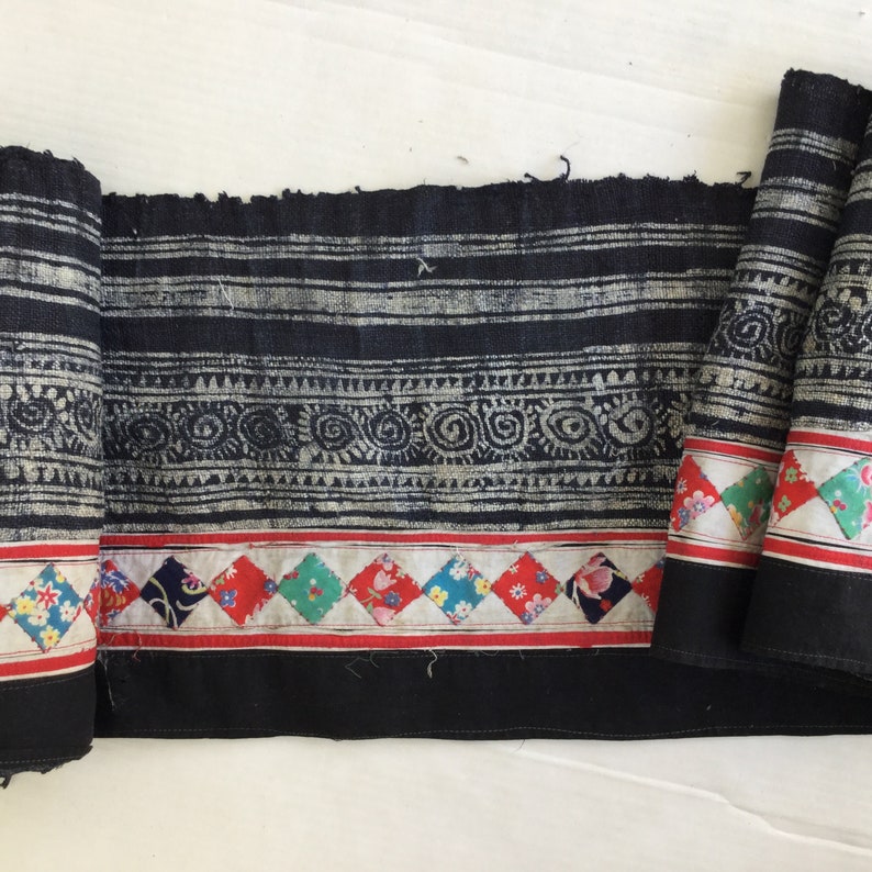 May include: A rolled-up length of indigo fabric with a detailed pattern of white stripes, circles, and triangles. The bottom edge features a row of colorful diamond-shaped patches with floral designs, bordered by red and black stripes.