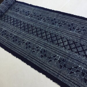 Puede incluir: Un rollo de tela teñida con índigo con un patrón geométrico. El diseño presenta filas de diamantes, triángulos y líneas rectas en blanco sobre un fondo azul oscuro. La tela tiene un borde en bruto deshilachado.