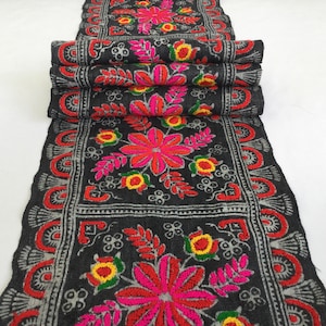 May include: Black table runner with a floral embroidery design. The embroidery features pink, red, yellow, and green flowers on a black background. The runner has a silver-grey border with a repeating pattern.