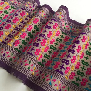 Pode incluir: Close-up de um tecido dobrado com fundo bege e um padrão repetitivo. O padrão apresenta formas bordadas coloridas em rosa, amarelo, verde e roxo. As bordas são aparadas com uma borda roxa escura.