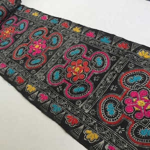 May include: Black fabric trim with colorful embroidered geometric patterns. The trim features a repeating design of four-leaf clover shapes with intricate details.