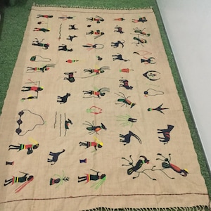 May include: A rectangular beige textile wall hanging featuring a repeating pattern of embroidered figures, animals, and insects in black, red, yellow, and green thread. The fabric has a fringed edge at the top and bottom, with a simple stitched border.