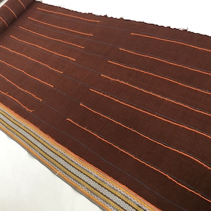 Può includere: Un tessuto marrone con strisce arancioni. Il tessuto ha un bordo decorativo con strisce gialle, arancioni e marroni.