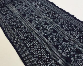 May include: A roll of indigo-dyed fabric with a repeating white geometric pattern. The design includes stripes, swirls, and diamond shapes. The fabric has a fringed edge and is likely used for textiles or home decor.