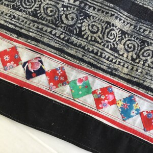 May include: Close-up of a textile with a dark blue and white patterned design. A white strip with red borders and colorful square patches with floral patterns is sewn onto the fabric. A black border finishes the edge.
