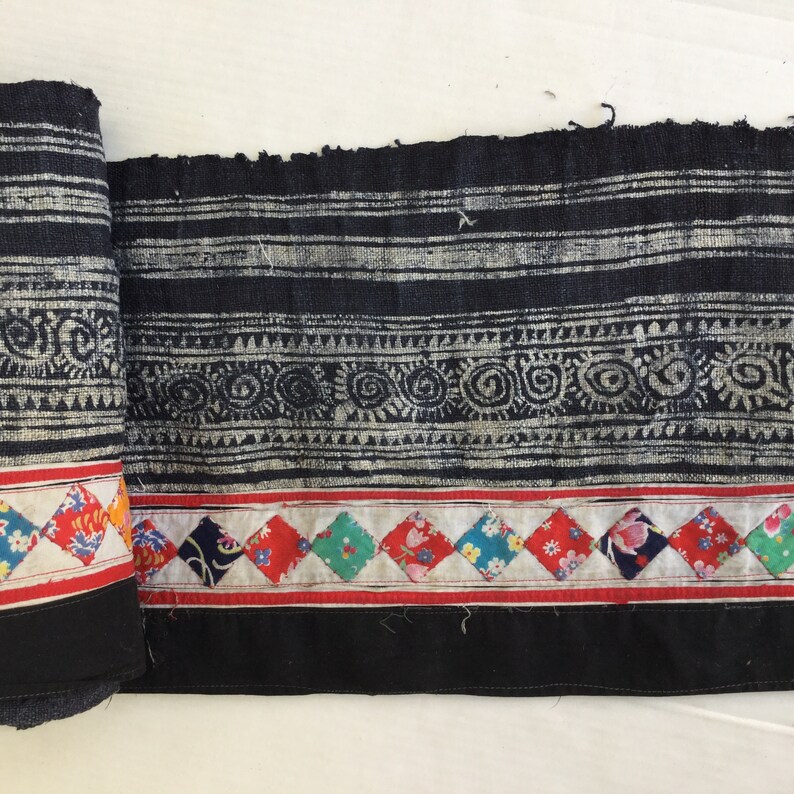 May include: Rolled textile with a black and white patterned design. The fabric features horizontal stripes, geometric shapes, and floral diamond patterns in red, white, and various colors. The edges are finished with a black border.