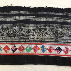 May include: Close-up of a textile fragment with a black and silver striped pattern at the top, followed by a decorative swirl design. Below, a row of colorful diamond-shaped patches with floral prints and a red stripe. The bottom is solid black.