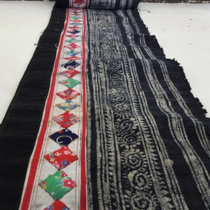 May include: A length of black fabric with a central patterned section and a decorative border. The border features a red stripe and diamond-shaped patches with floral and geometric designs. The patterned section has a white and black design.