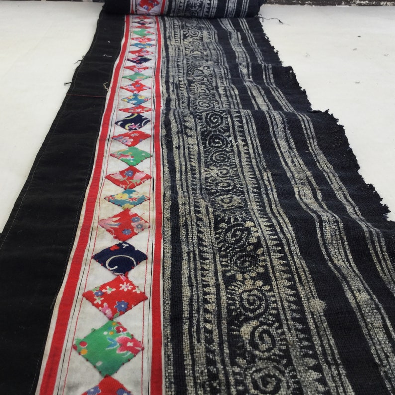 May include: A length of black fabric with a central patterned section and a decorative border. The border features a red stripe and diamond-shaped patches with floral and geometric designs. The patterned section has a white and black design.