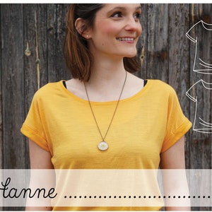 May include: A woman wearing a yellow t-shirt with a round pendant necklace. The t-shirt has rolled up sleeves. The background is a wooden wall with two line drawings of t-shirts.