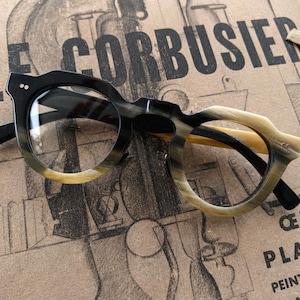May include: Black and tan-colored eyeglasses with a unique frame design, resting on a vintage book. The glasses have a rounded shape with a gradient color transition. The book's cover features the text "LE CORBUSIER".