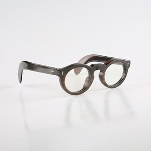 Puede incluir: Gafas de montura redonda con efecto veteado marrón oscuro. Las gafas tienen patillas gruesas y curvadas y un puente de cerradura. Las lentes son transparentes y la montura presenta pequeños remaches decorativos cerca de las bisagras.