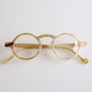 Può includere: Occhiali rotondi con montatura in corno di colore marrone chiaro e crema. Gli occhiali hanno uno stile classico e vintage.