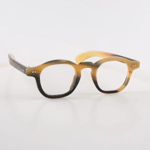 Può includere: Un paio di occhiali con montatura rotonda. La montatura è un mix di marrone scuro e beige. Le aste sono marrone scuro e le lenti sono trasparenti. Gli occhiali hanno uno stile classico e vintage.