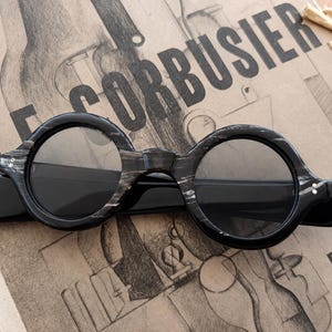 May include: Round-framed sunglasses with a black and gray marbled pattern. The sunglasses have dark lenses and a thick frame. The text "LE CORBUSIER" is visible in the background.