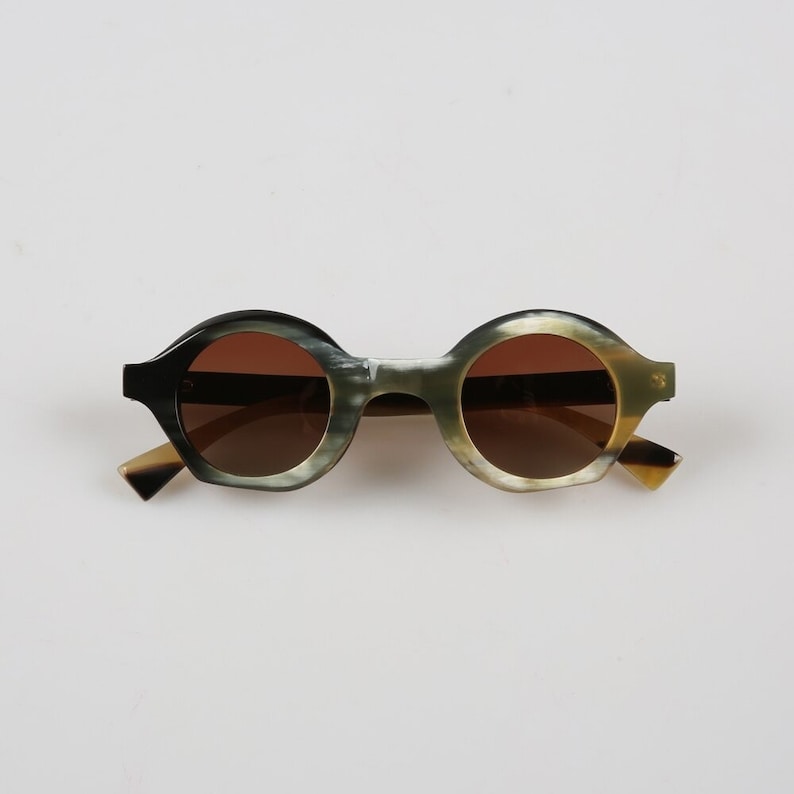 May include: Round tortoiseshell sunglasses with brown lenses. The sunglasses have a black and green pattern on the frames.