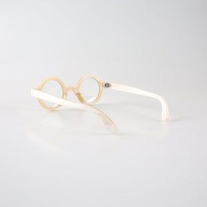 K&ouml;nnte beinhalten: Eine runde, cremefarbene Brille mit wei&szlig;em Rahmen. Die Brille liegt auf einem wei&szlig;en Hintergrund.