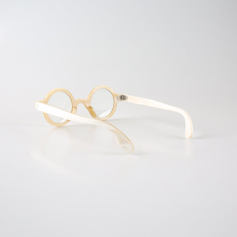 K&ouml;nnte beinhalten: Eine runde, cremefarbene Brille mit wei&szlig;em Rahmen. Die Brille liegt auf einem wei&szlig;en Hintergrund.